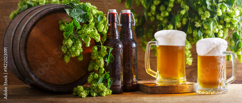 mugs of beer, green hops, bottles and beer barrel on wooden background