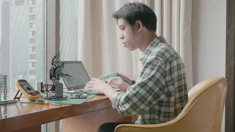 A man is sitting on a couch and working on a project. He is using a laptop and a small robot. Scene is focused and determined A man is sitting on a couch and working on a project. He is using a laptop and a small robot. Scene is focused and determined