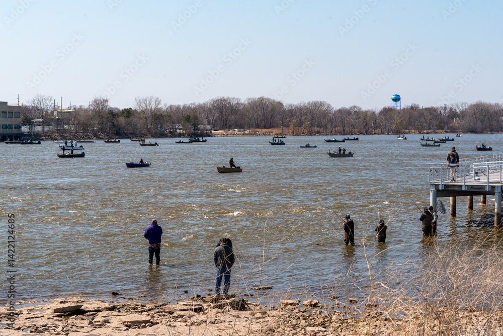 People fishing in boats and on the river shoreline on Fox river in Spring at De Pere, Wisconsin