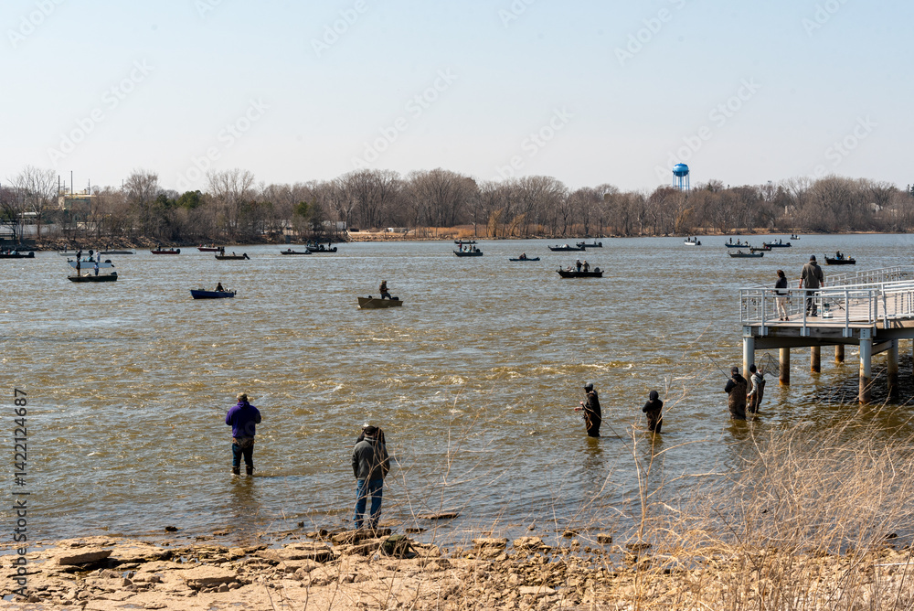 People fishing in boats and on the river shoreline on Fox river in Spring at De Pere, Wisconsin