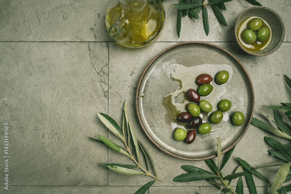 Green and black olives in a bowls and olive oil on gray background top view. Olive oil and variety of green and black olives in bowls with olive leaves all around taken from above.