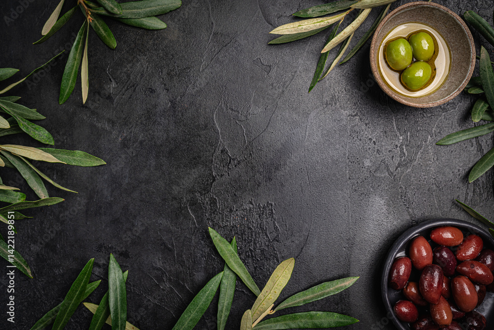 Green and black olives in a bowls and olive oil on dark background top view. Olive oil and variety of green and black olives in bowls with olive leaves all around taken from above.