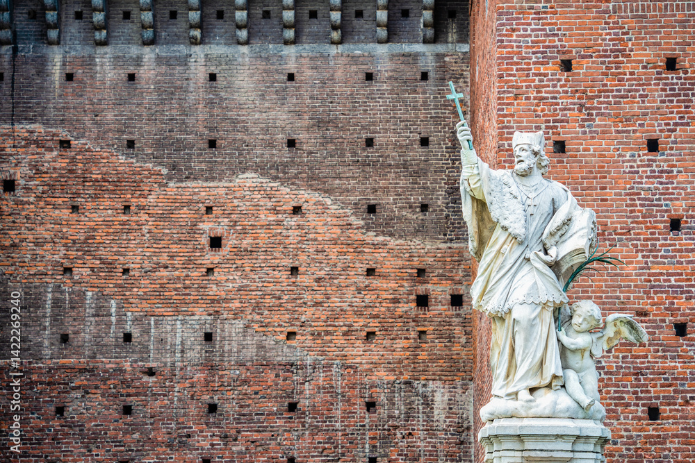 Naklejka premium Marble statue of a saint holding a cross in front of the historic brick walls of Castello Sforzesco in Milan, Italy