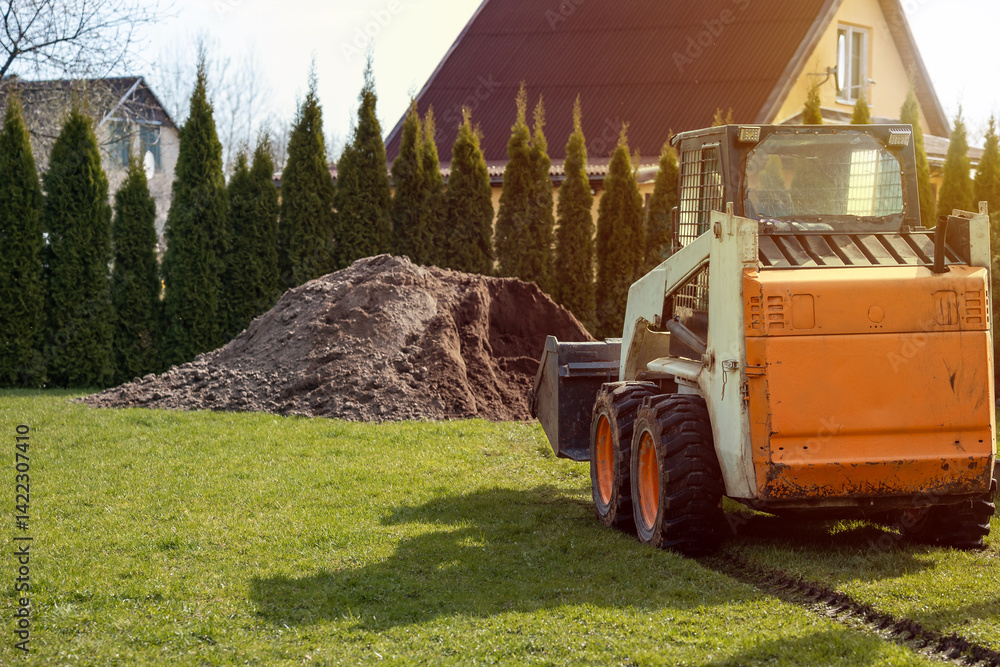 An orange and white skid steer loader sits on grass with tire tracks nearby. A soil pile and tall evergreen trees partially obscure a yellow house.
