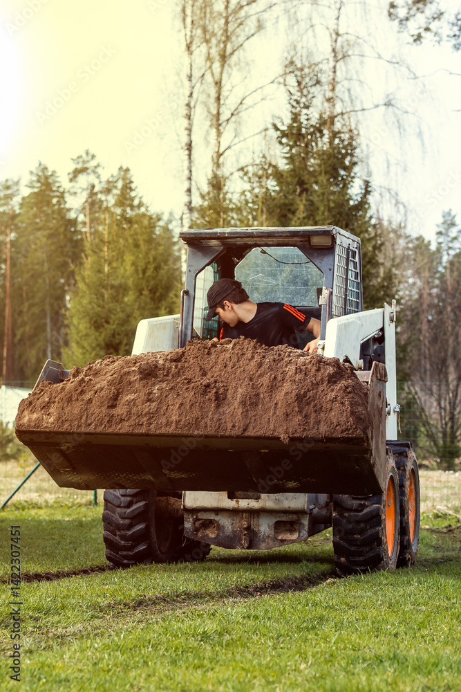 A man in a dark shirt and cap operates a white skid steer loader with orange wheels, carrying soil in the bucket. Tall trees, including conifers and birches, are visible.