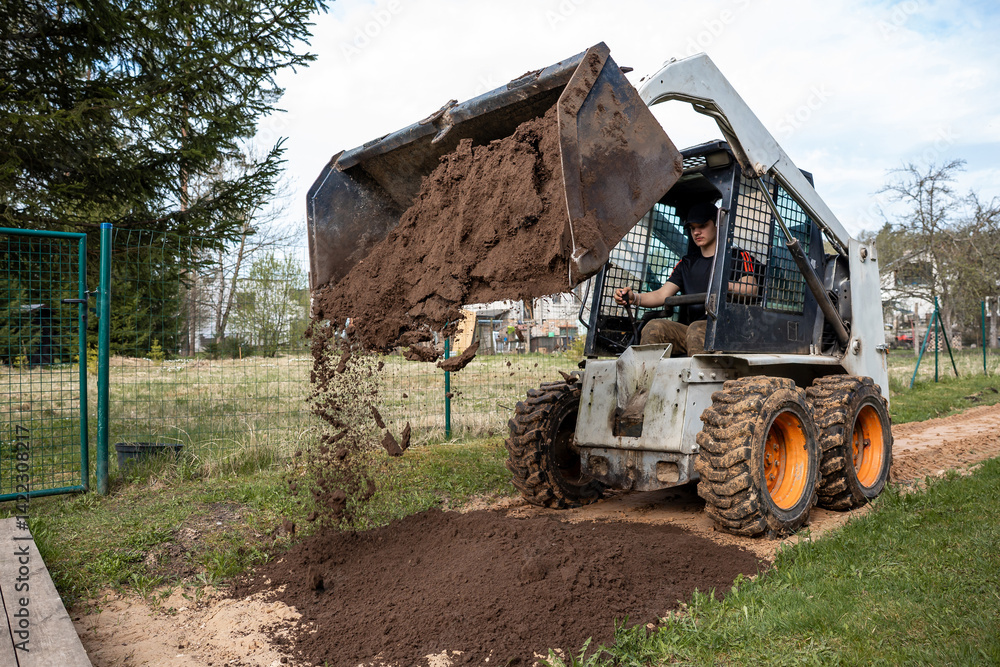 A skid steer loader with orange wheels dumps soil onto a pathway. The operator works in a fenced area with grass, sparse trees, and distant buildings.