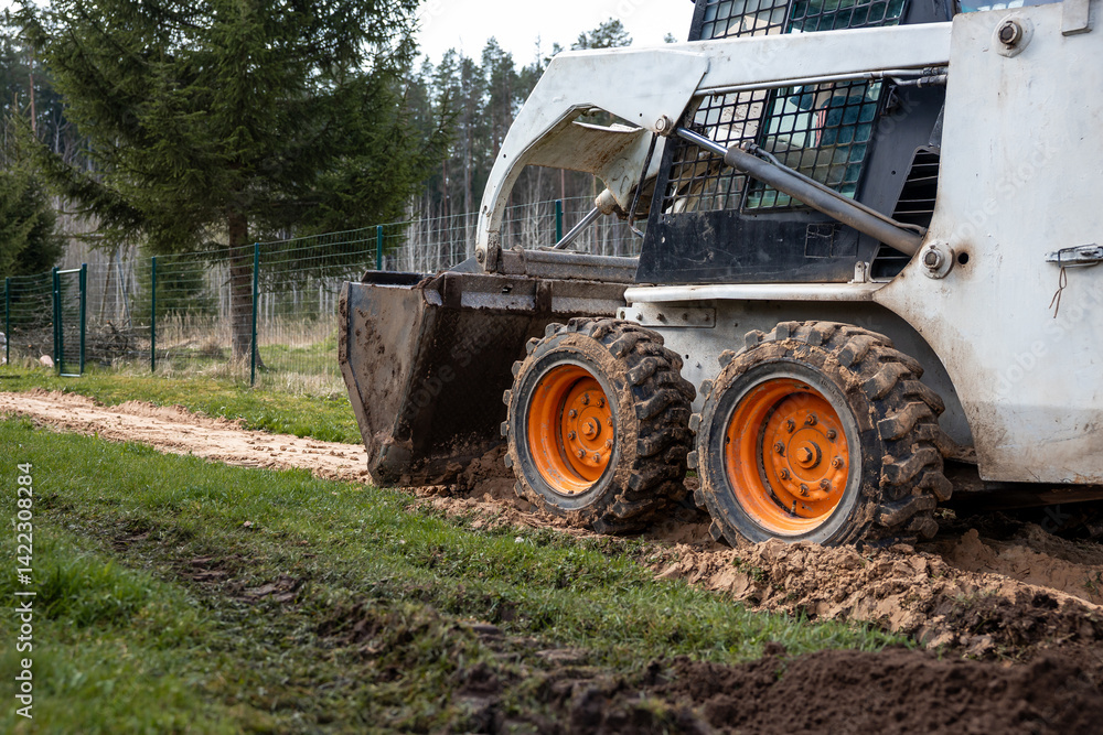 A skid steer loader with orange wheels works on a dirt path, leaving tire tracks. The bucket is lowered, with a wire fence and evergreen trees nearby.