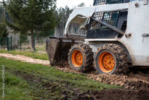 Obraz na płótnie A skid steer loader with orange wheels works on a dirt path, leaving tire tracks. The bucket is lowered, with a wire fence and evergreen trees nearby. w salonie