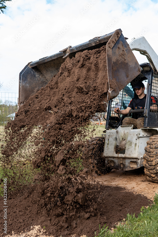 A worker uses a skid steer loader to dump loose soil onto the ground. The scene includes a wire fence, sparse vegetation, and a partly cloudy sky.