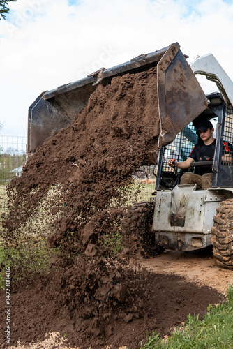 Obraz na płótnie A worker uses a skid steer loader to dump loose soil onto the ground. The scene includes a wire fence, sparse vegetation, and a partly cloudy sky. w salonie