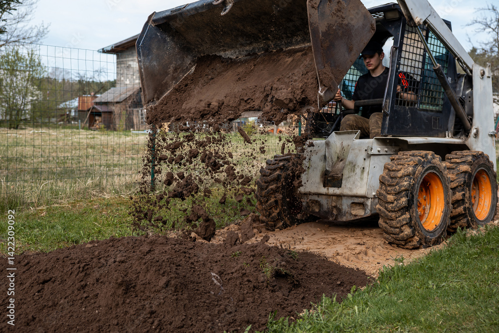 A skid steer loader with orange wheels dumps soil onto the ground, scattering dirt mid air. A wire fence and wooden structures are visible nearby.