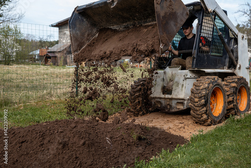 Obraz na płótnie A skid steer loader with orange wheels dumps soil onto the ground, scattering dirt mid air. A wire fence and wooden structures are visible nearby. w salonie