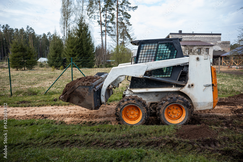 A skid steer loader with orange wheels transports soil on a dirt path. The area is fenced, with evergreen trees and a modern flat roof building visible.