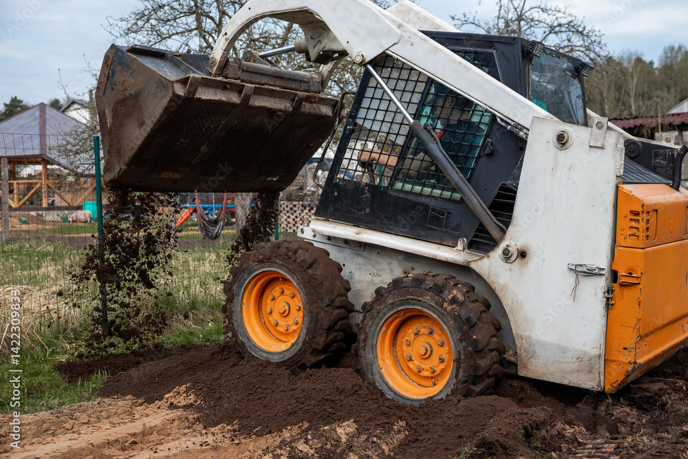 A white and orange skid steer loader with raised bucket dumps soil on uneven ground. Nearby are a fenced area, playground equipment, and sparse trees.