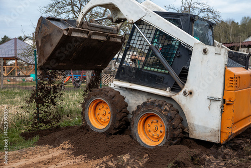 Obraz na płótnie A white and orange skid steer loader with raised bucket dumps soil on uneven ground. Nearby are a fenced area, playground equipment, and sparse trees. w salonie