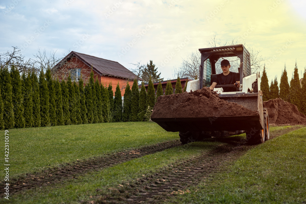 A man drives a small loader carrying soil across a grassy area with visible tire tracks. A wooden house and tall evergreen trees are in the scene.