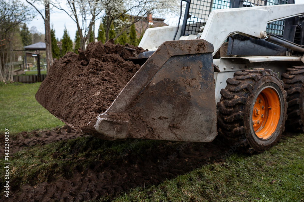 A white skid steer loader with orange wheels and a soil filled bucket on grass. Tire tracks, trees, a gazebo, and a building are visible nearby.