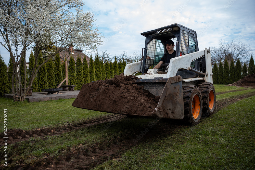 A skid steer loader moves soil on grass, leaving tire tracks. A blossoming tree, trimmed hedges, a wooden platform, and a house roof are visible.