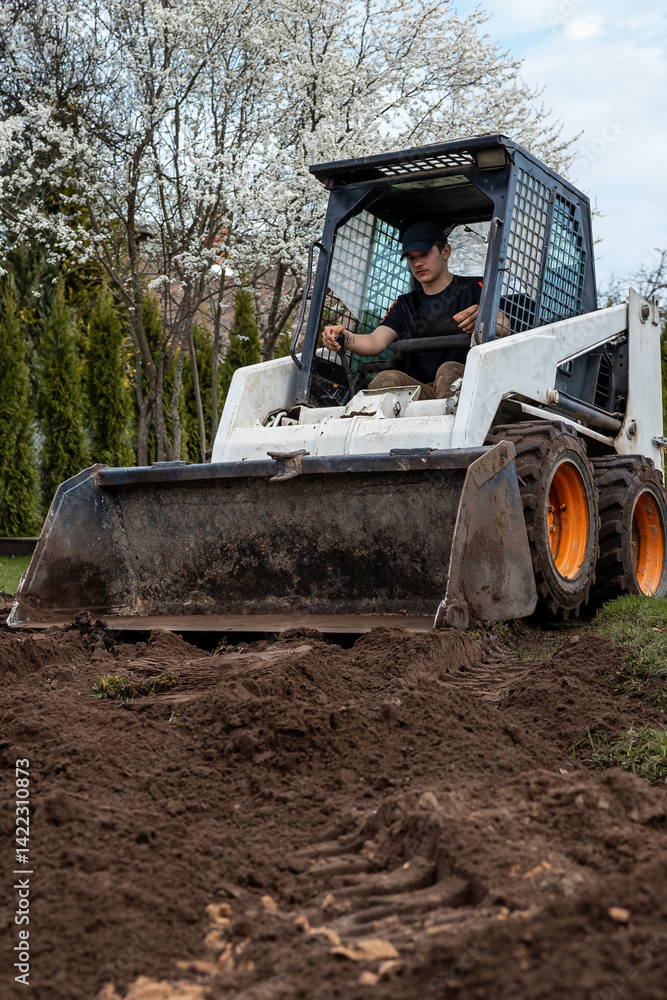A man uses a skid steer loader with orange wheels and a front bucket to move soil. Flowering trees with white blossoms and hedges are visible behind.