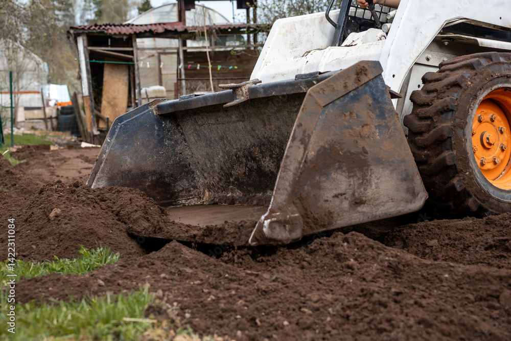 A small construction vehicle with an orange wheel and front loader moves soil near a rustic structure with a corrugated metal roof and wooden panels.
