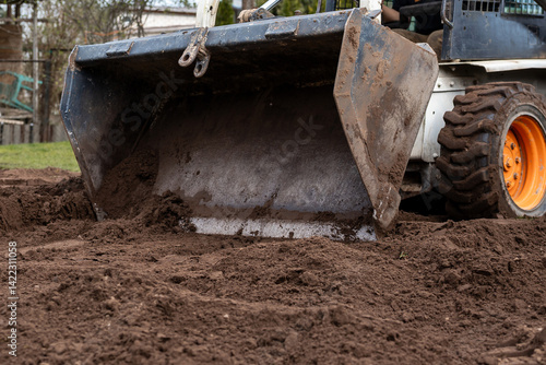 Obraz na płótnie Close up of a white construction vehicle with orange wheels and a metal bucket moving soil. Dirt and mud on the bucket indicate recent use. w salonie