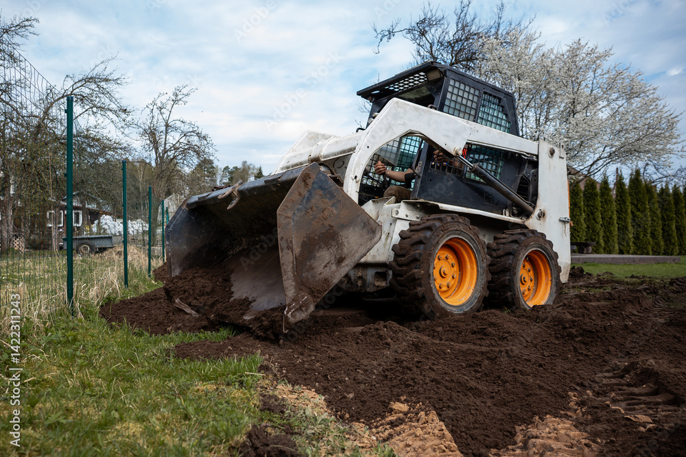 A skid steer loader with orange wheels moves soil near a wire fence. Trees, some in bloom, and tall shrubs surround the outdoor work area.