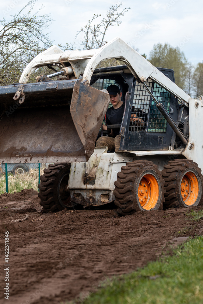 A skid steer loader with orange wheels carries soil in its raised bucket. A person operates the machine in a grassy field with sparse trees nearby.