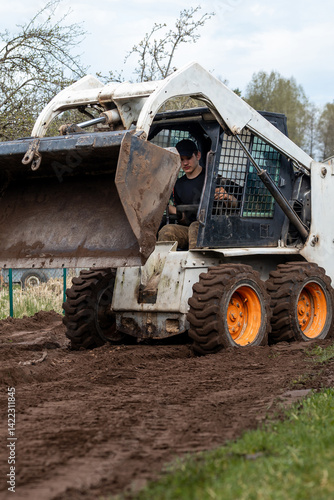 Obraz na płótnie A skid steer loader with orange wheels carries soil in its raised bucket. A person operates the machine in a grassy field with sparse trees nearby. w salonie