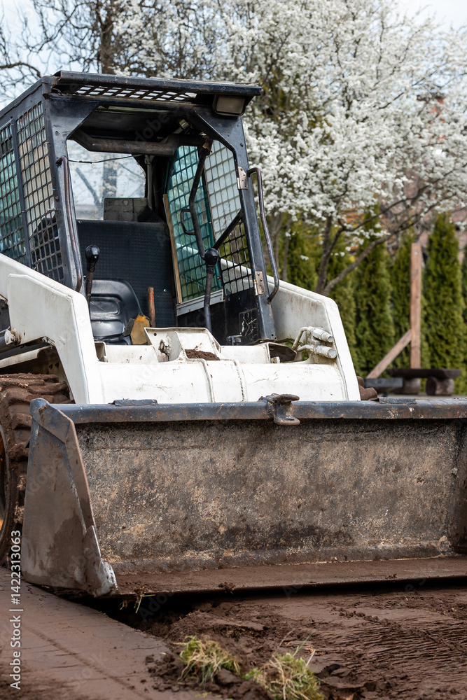 A white skid steer loader with a weathered bucket sits on dirt, surrounded by blooming white trees and trimmed coniferous hedges in spring.