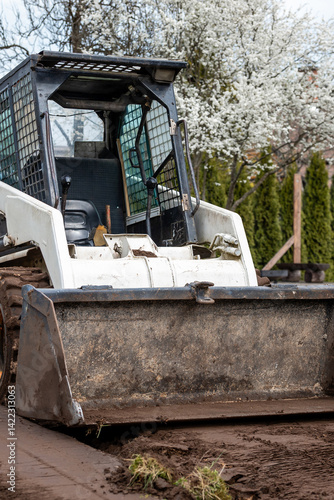A white skid steer loader with a weathered bucket sits on dirt, surrounded by blooming white trees and trimmed coniferous hedges in spring.
