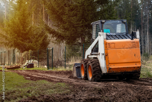 Obraz na płótnie An orange and white skid steer loader parked on a dirt path with visible tire tracks, bordered by a metal fence and surrounded by tall evergreen trees. w salonie