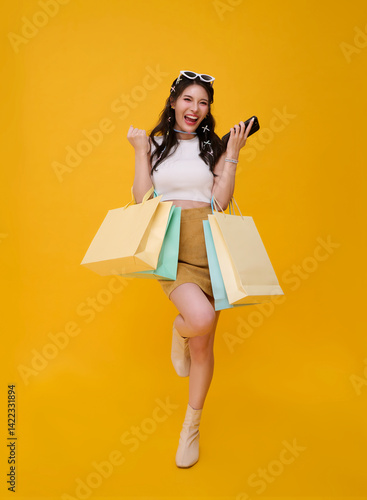 Happy celebrating cute Asian woman shopper carrying colorful shopping bags isolated on yellow background. shopping discount promotion concept.