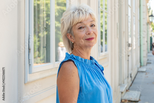 A stylish woman aged 65-70 in a blue outfit stands outside a building with window