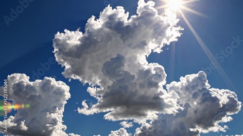 A white Crown-shaped cloud with a puffy and fluffy cumulus cloudscape is set against a sunny clear blue sky background with a lens flare effect or bright sunlight rays and sunbeams in a 4k