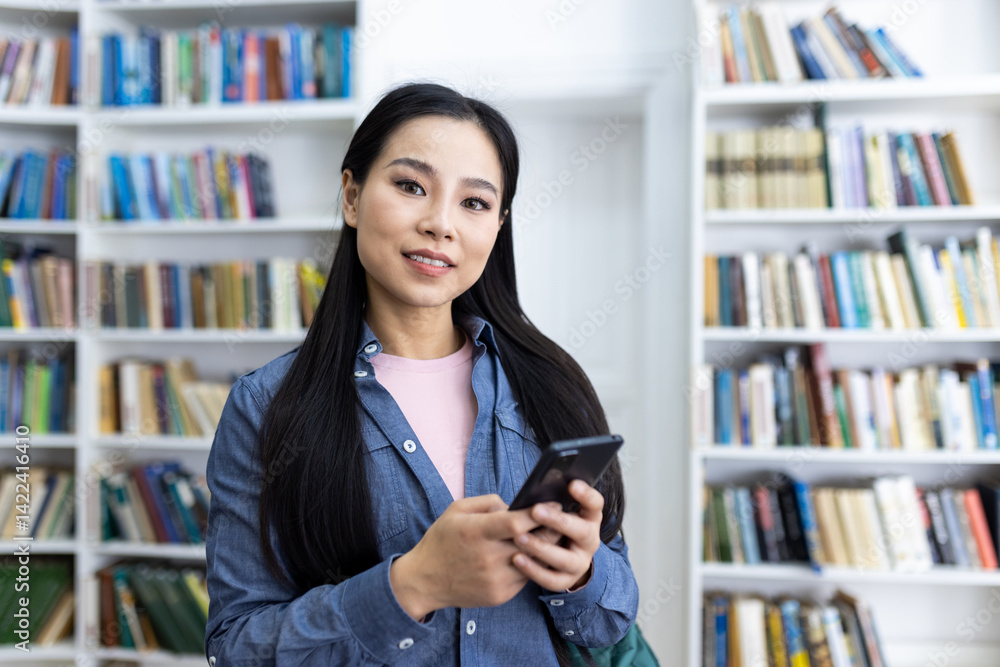 A smiling asian woman holding a smartphone in a library setting, surrounded by books and shelves. She is likely a student or a reader.