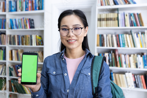 A young woman, wearing glasses and a backpack, presents a smartphone with a green screen in a library. Bookshelves line the background.