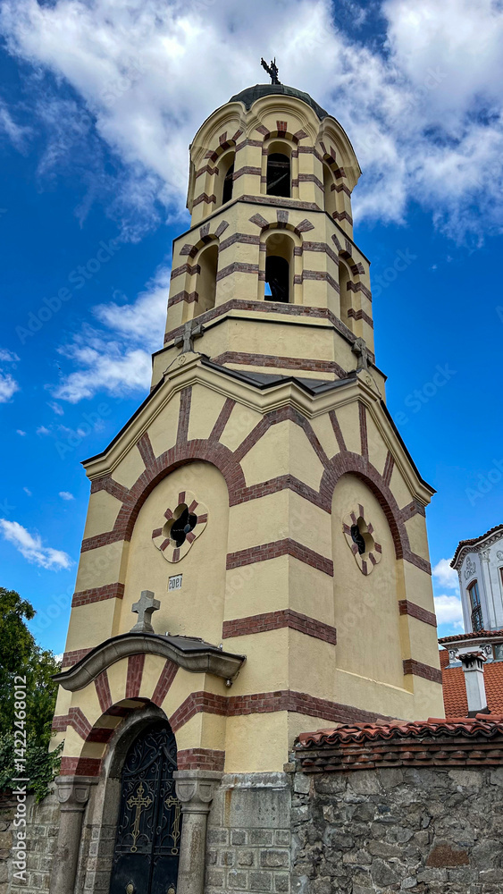 Fototapeta premium Historic Orthodox Church Bell Tower Built In 1905 With Cream And Red Brick Façade, Cross On Top, Arched Windows, Decorative Black Iron Gate, Stone Base, And Dramatic Sky Background