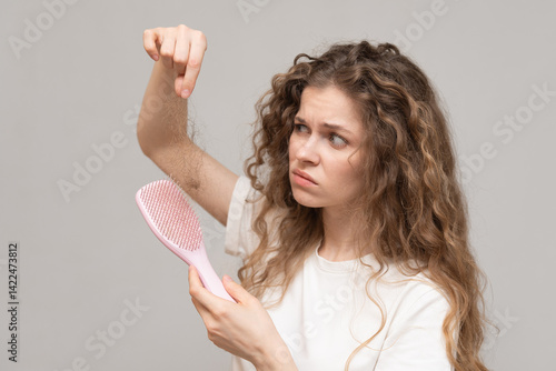 Frustrated young woman with curly hair inspecting hair loss from a pink hairbrush against a gray background.