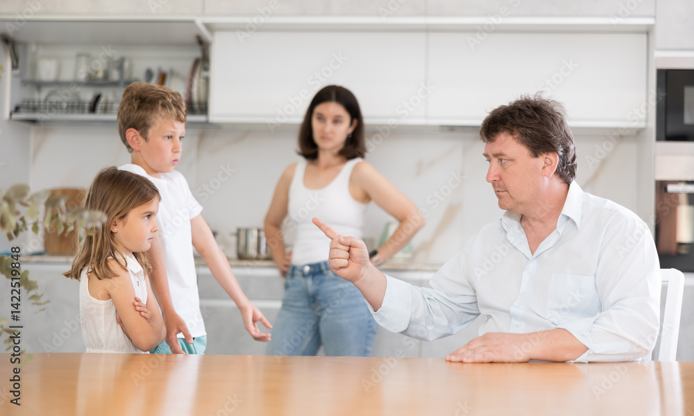 Fototapeta premium At home in kitchen, mom and dad scold young children for imprudent act. Husband and wife scold children, son denies his bad behavior. Children with guilty look stand in front of angry father