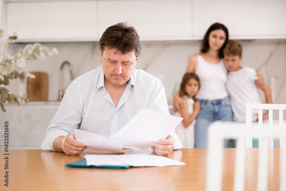 Upset father reads and looks through financial documents. Sad wife with children in the background