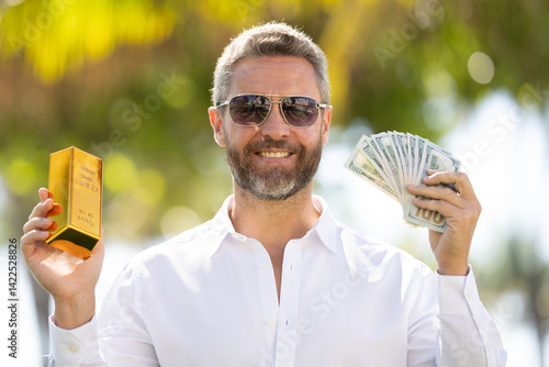 Rich businessman holding a gold bar and cash. Gold financial success. Excited rich man holding gold and money. Business man showcasing gold bars. Golden treasure. Hands hold golden bars and cash.
