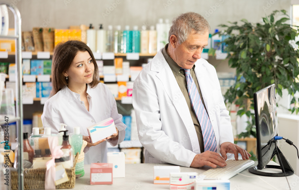 Elderly experienced pharmacist works at computer in a drugstore. Young assistant helps to find pills Elderly experienced pharmacist works at computer in a drugstore. Young assistant helps to find pills