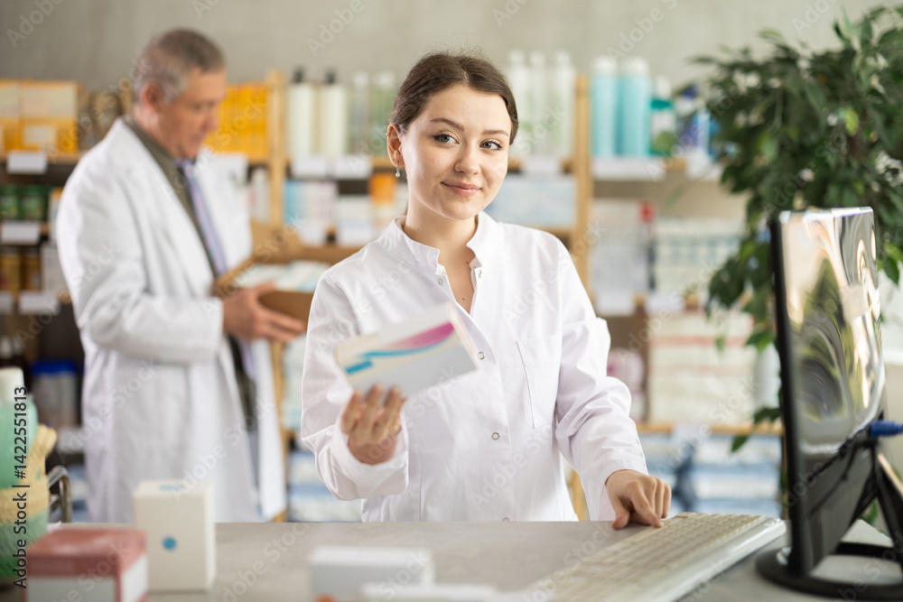 Obraz premium Girl pharmacist uses computer at workplace. Employee reads regulations and laws, makes report, reads information about drugs. Male colleague work in background