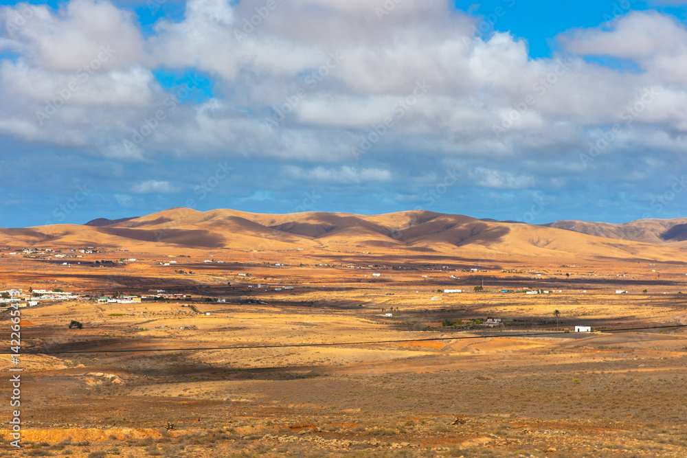 Golden hills stretch across the arid landscape of Fuerteventura under a dramatic sky. Scattered white houses dot the terrain, creating a striking contrast with the vast, sunlit desert