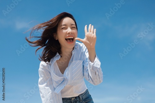 A cheerful Asian woman enjoying a sunny day at the beach. She is wearing a casual shirt and jeans standing in front of the blue sky background