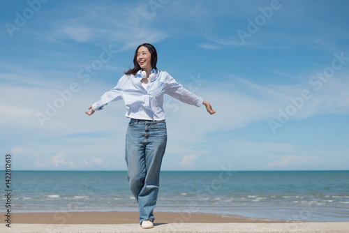 A cheerful Asian woman enjoying a sunny day at the beach. She is wearing a casual shirt and jeans with headphones around her neck, standing in front of the beach