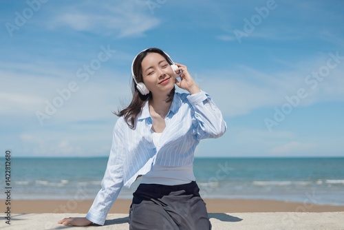 A joyful young Asian woman listening to music with headphones at the beach, smiling brightly under the clear blue sky.enjoying and relaxing summer atmosphere concept.