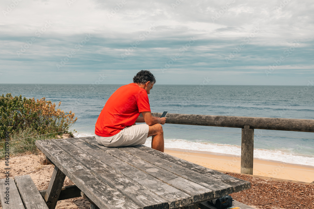 Man sitting on picnic bench at ocean lookout looking at his phone Man sitting on picnic bench at ocean lookout looking at his phone