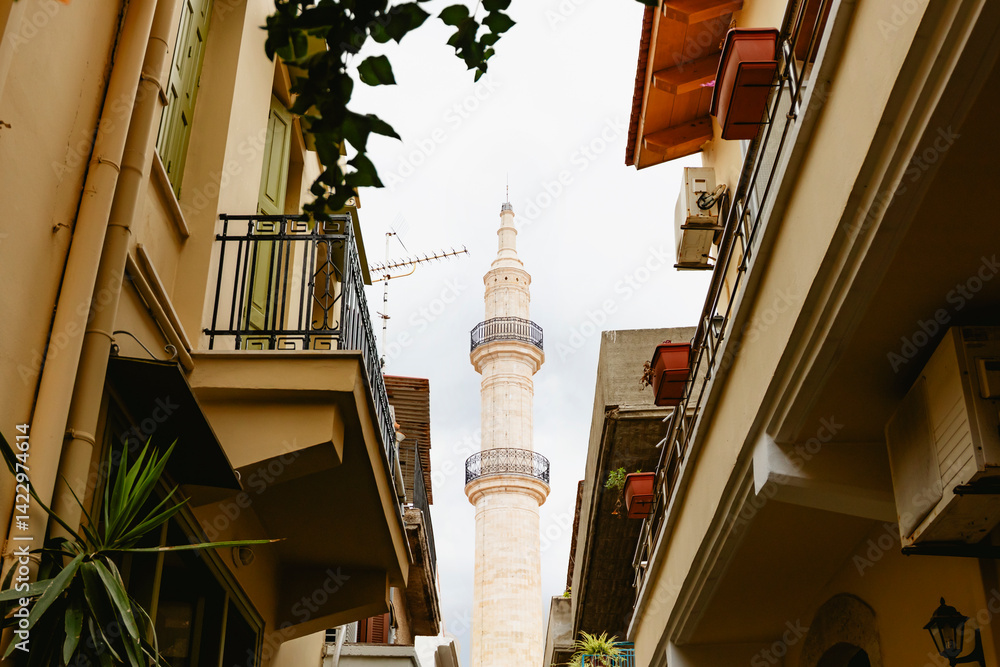 A delightful alley bordered by historic buildings leads to an impressive minaret towering beneath a cloudy sky. Rethymno town, Crete island, Greece.