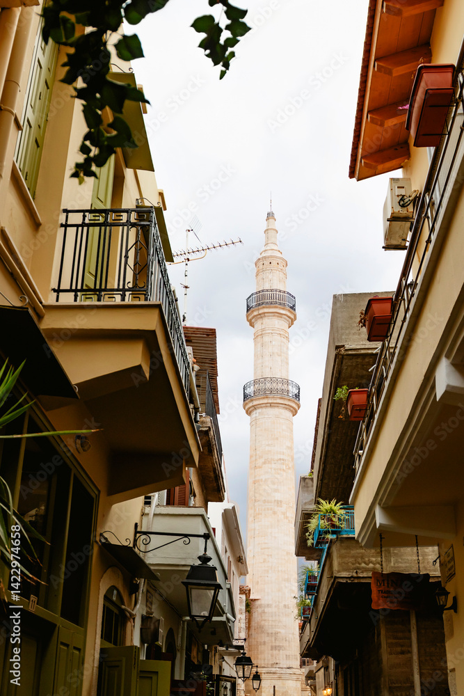 A delightful alley bordered by historic buildings leads to an impressive minaret towering beneath a cloudy sky. Rethymno town, Crete island, Greece.